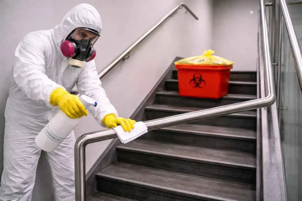 A single biohazard cleanup technician in full hazmat suit and respirator disinfecting a stairwell handrail while working in a clean indoor environment.